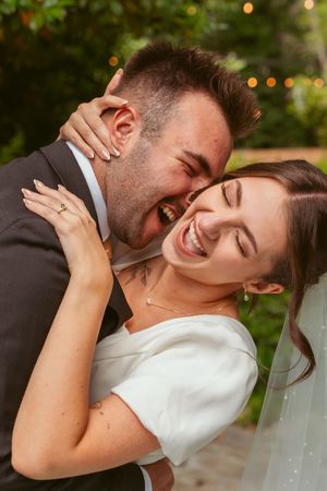 Two newlyweds sharing a joyful laugh together during their wedding day with outdoor bokeh lighting in the background.