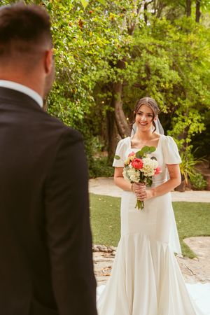 A bride in a white wedding dress with short sleeves holds a colorful bouquet while standing outdoors surrounded by greenery.