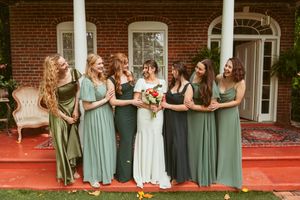 A wedding party wearing mint and sage green bridesmaid dresses laughs together on a red porch of a brick house.