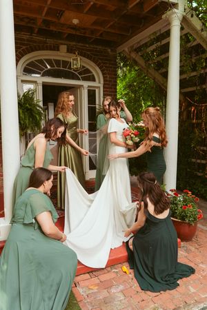 Wedding party in sage green dresses helps adjust a flowing white bridal gown on a brick porch with white columns.