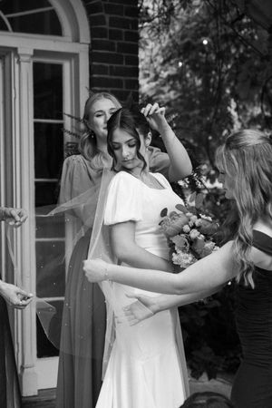 Black and white photo showing preparations for a wedding celebration, with bridal party and flowers outside a brick building.