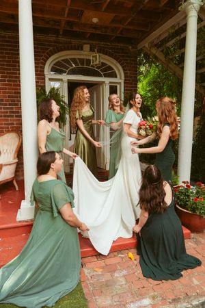 Wedding party in sage green dresses helps adjust the bride's gown on a brick porch with white columns.