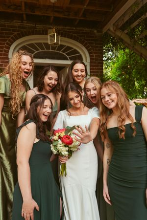 Bridesmaids in dark green dresses gather around bride looking at phone and laughing under brick archway.