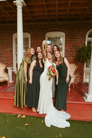 Wedding party in coordinating green dresses and white gown pose on brick porch with red carpet.