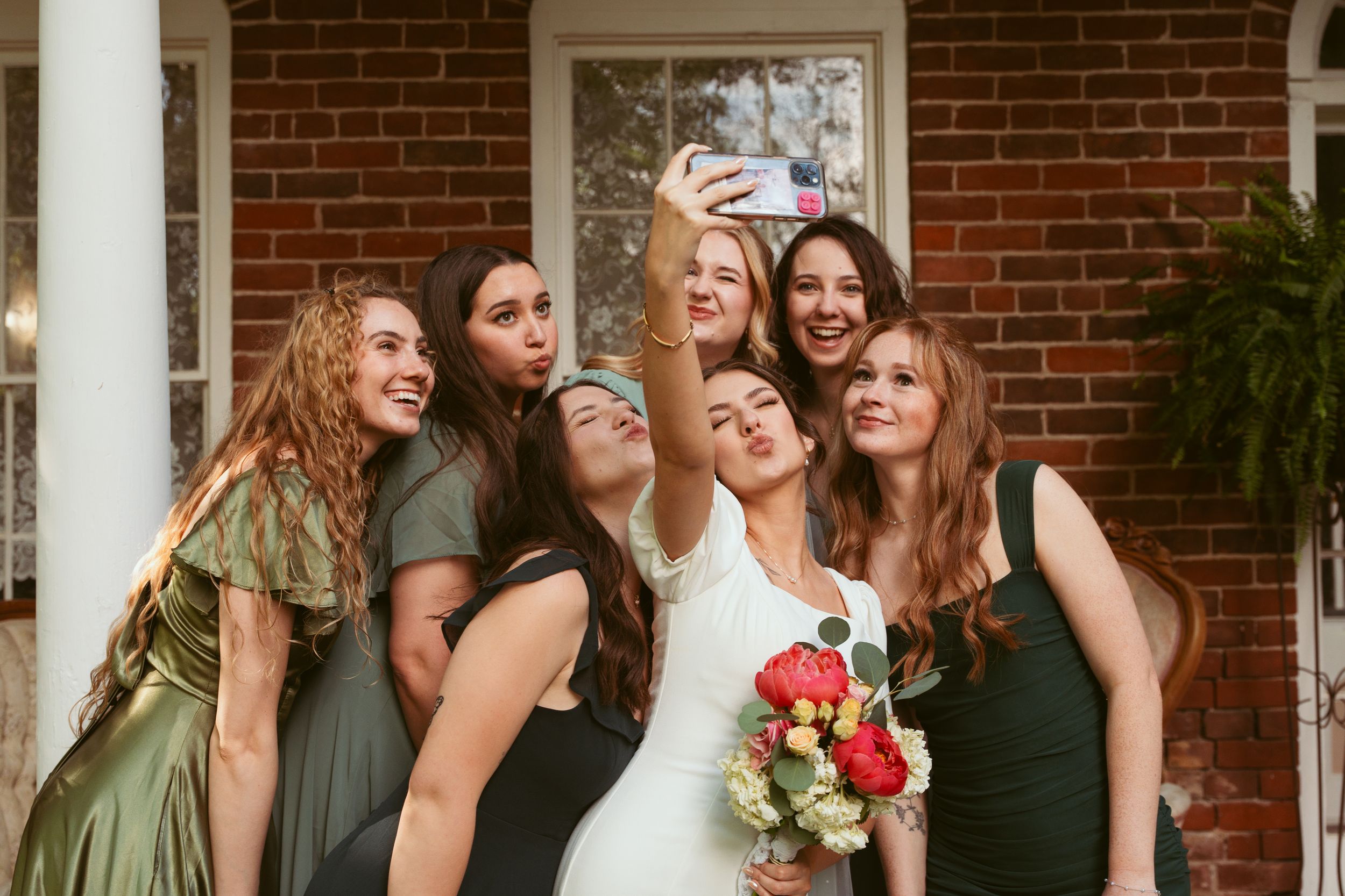 Wedding party takes joyful selfie together on brick porch during celebration.