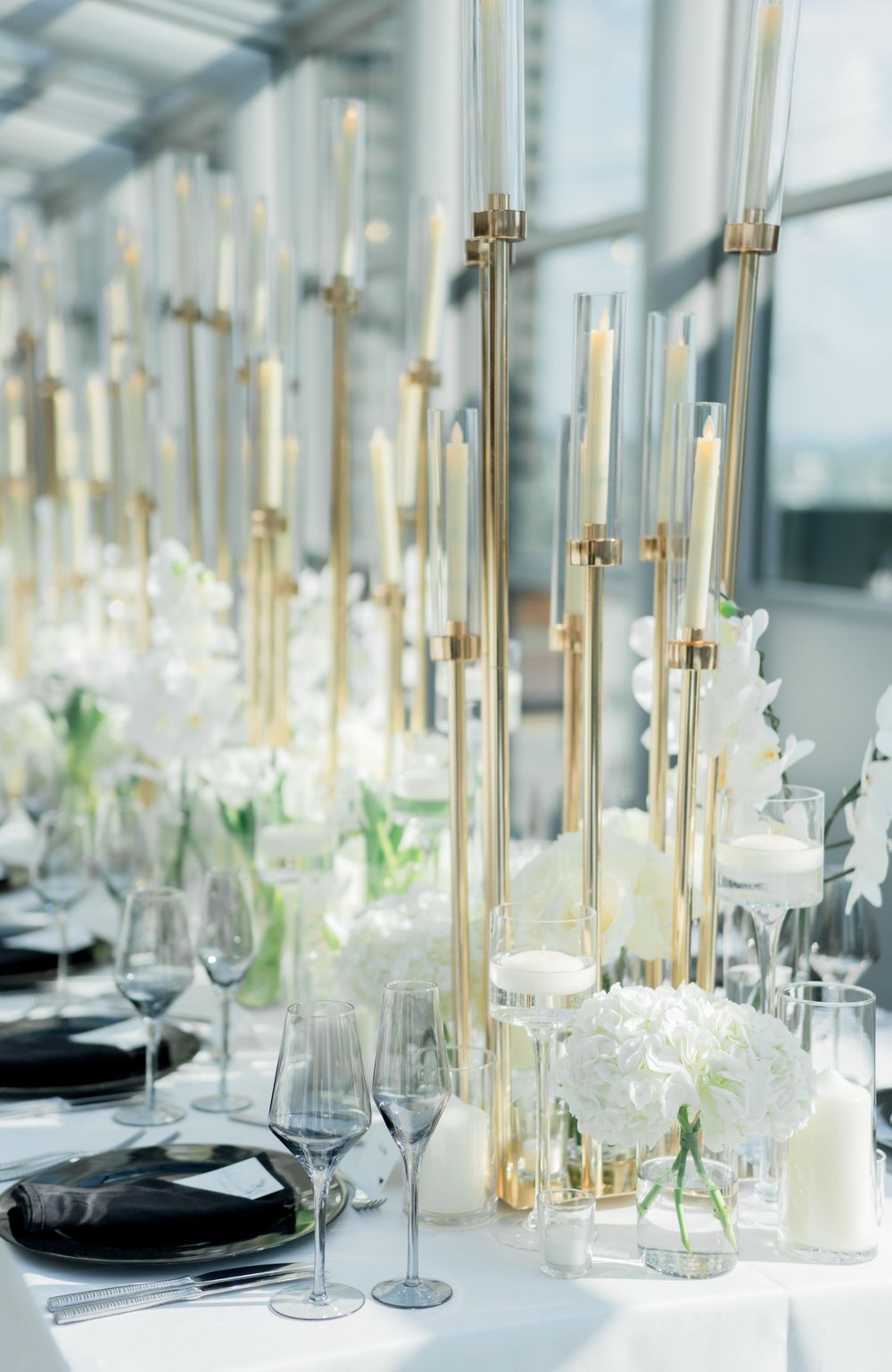 Elegant wedding table setting with tall gold candlesticks, white hydrangeas, and crystal glassware against a bright window.