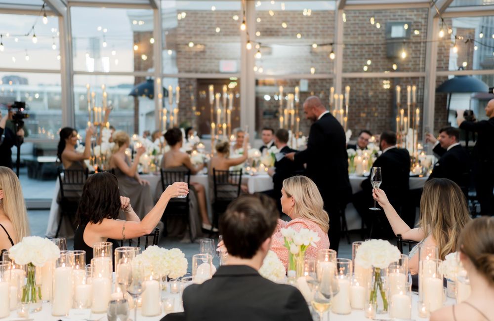 Wedding reception guests seated at elegant long tables with glowing candlelight and floral centerpieces in a bright venue.