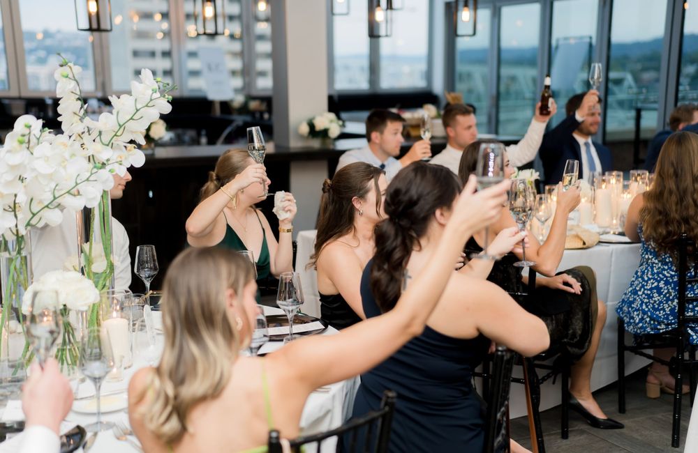 Elegant wedding guests in black dresses raise their glasses for a toast at a reception with white floral centerpieces.