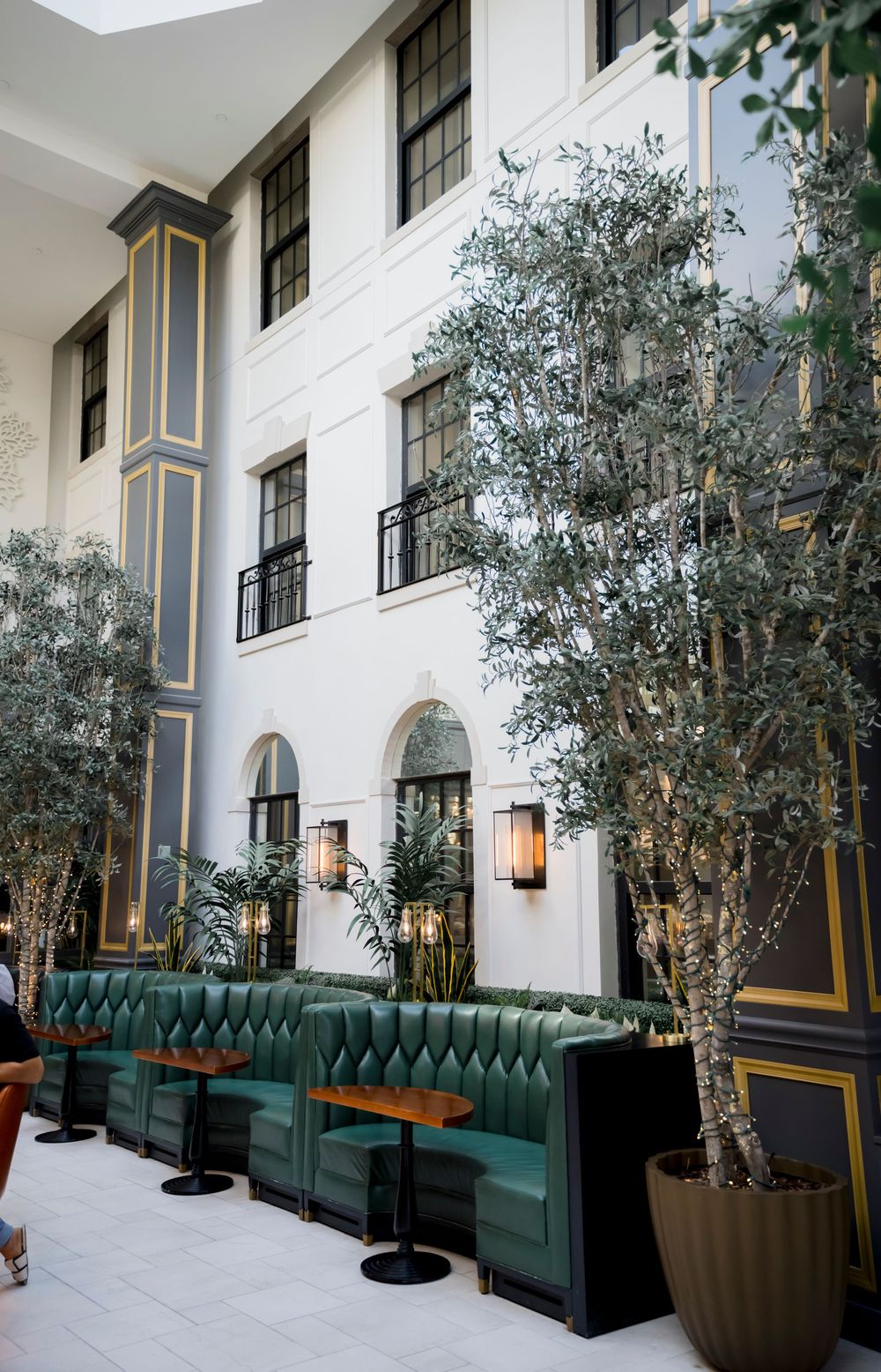 Elegant hotel interior with green velvet banquettes and olive trees against a white architectural facade.