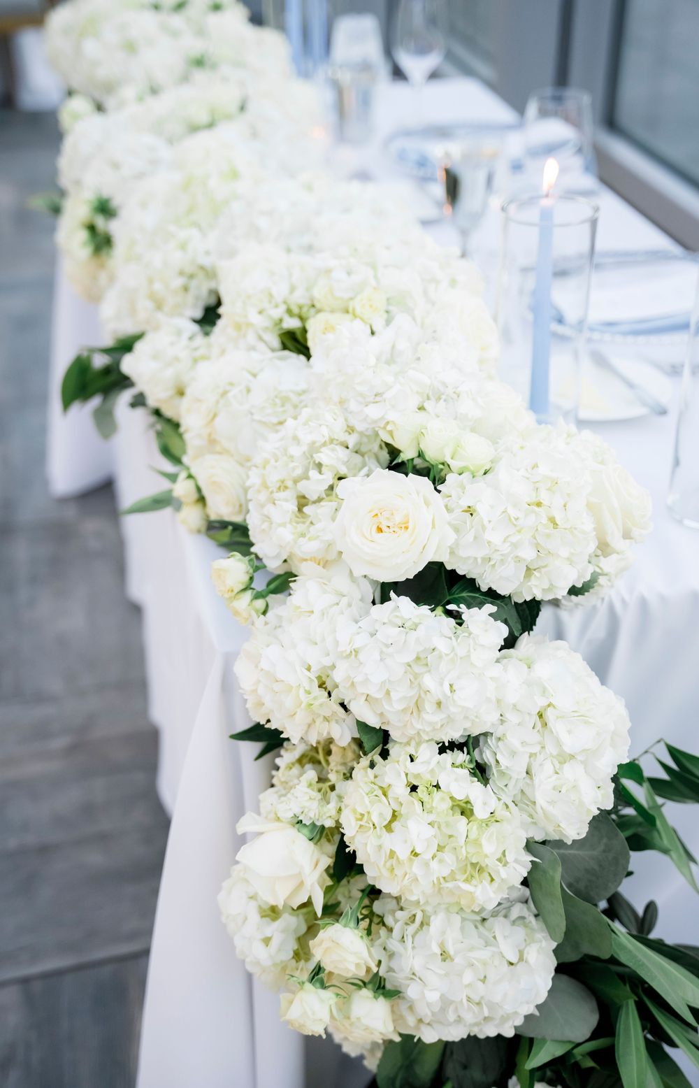 Elegant white hydrangeas and roses arranged in a cascading floral runner for a wedding reception table.