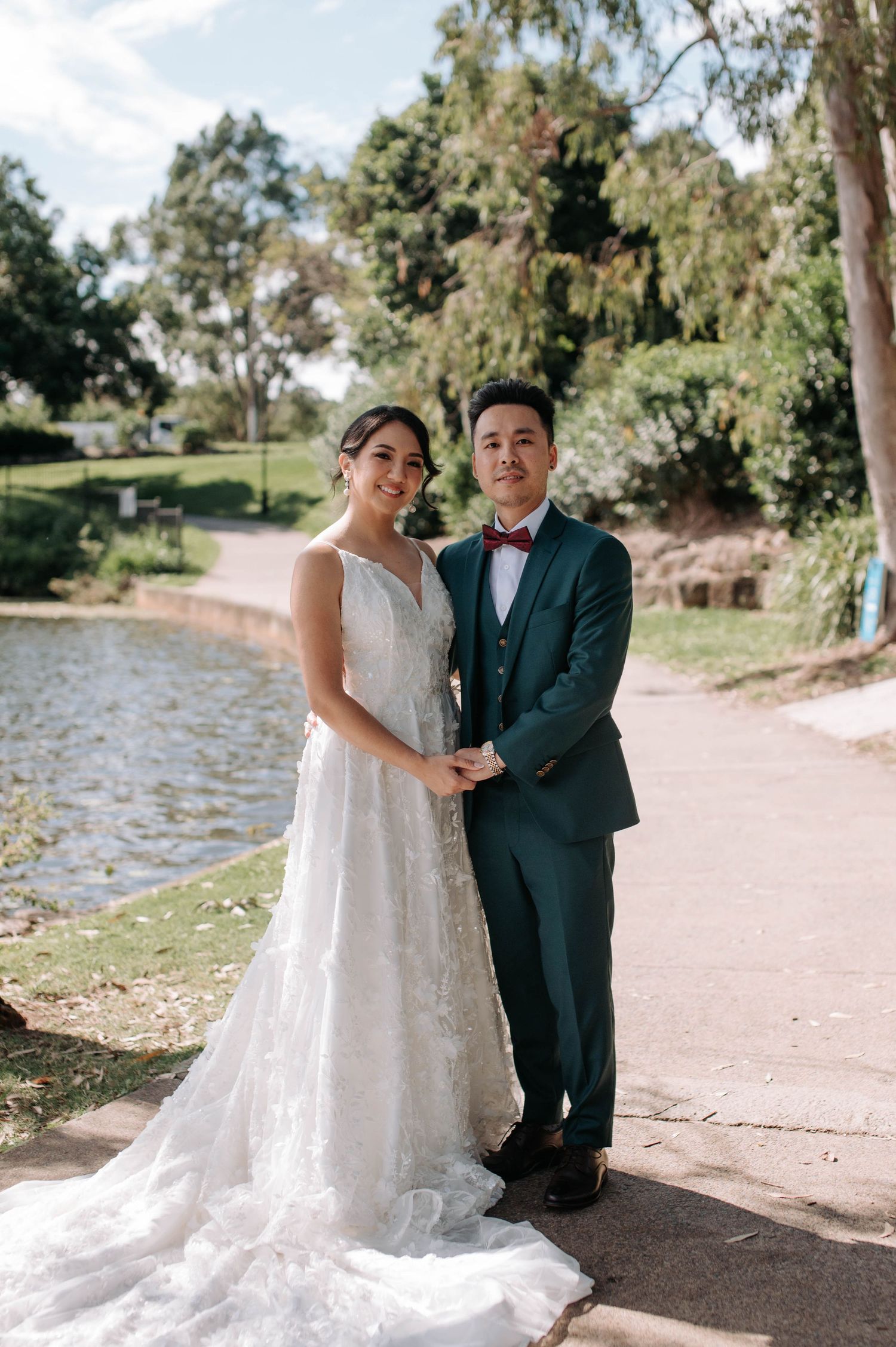 A wedding couple stands on a lakeside path surrounded by trees on a sunny afternoon.