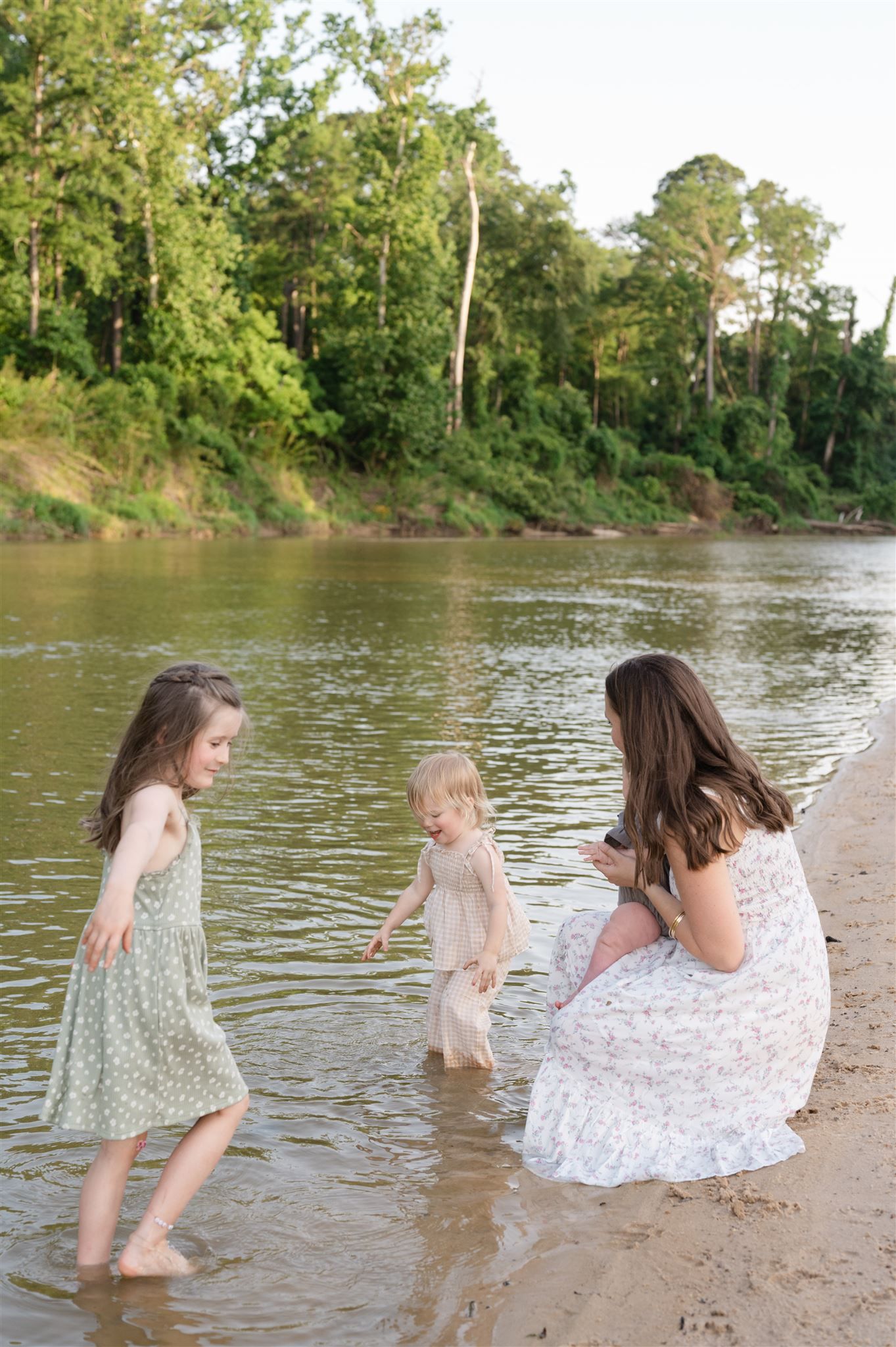 People in flowing dresses playing at the water's edge of a serene river surrounded by green trees.
