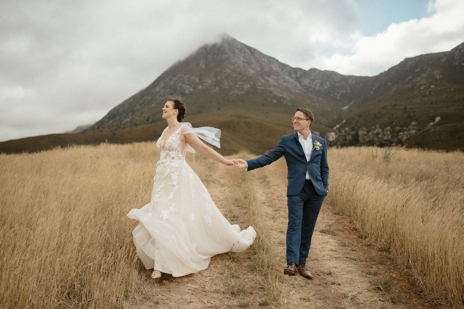 A couple holds hands while walking through tall golden grass with rugged mountain peaks in the background.