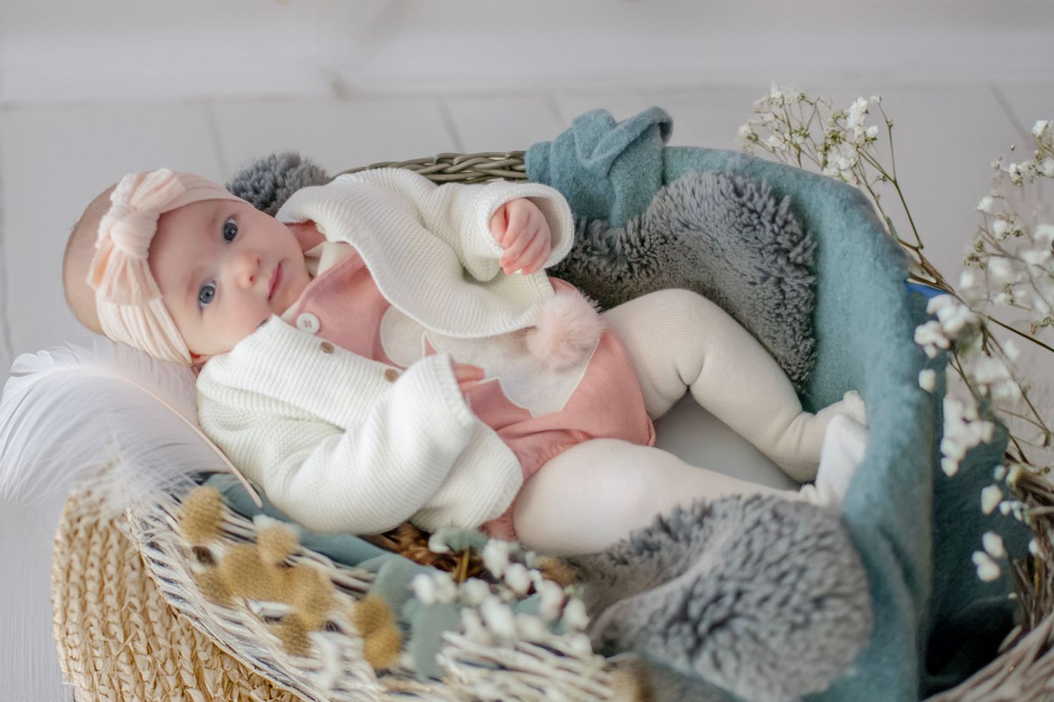 A baby in a white sweater lies in a cozy woven basket with floral decorations and gray blankets.