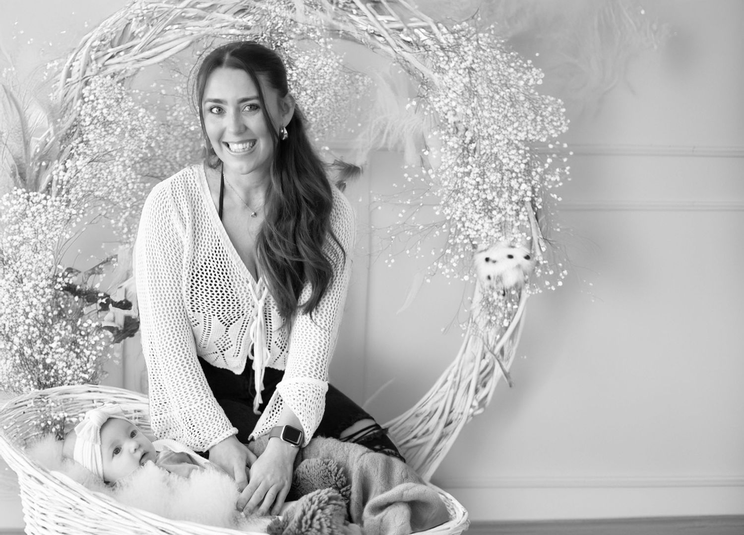 A black and white portrait with someone sitting by a decorative white floral wreath arrangement against a light wall.
