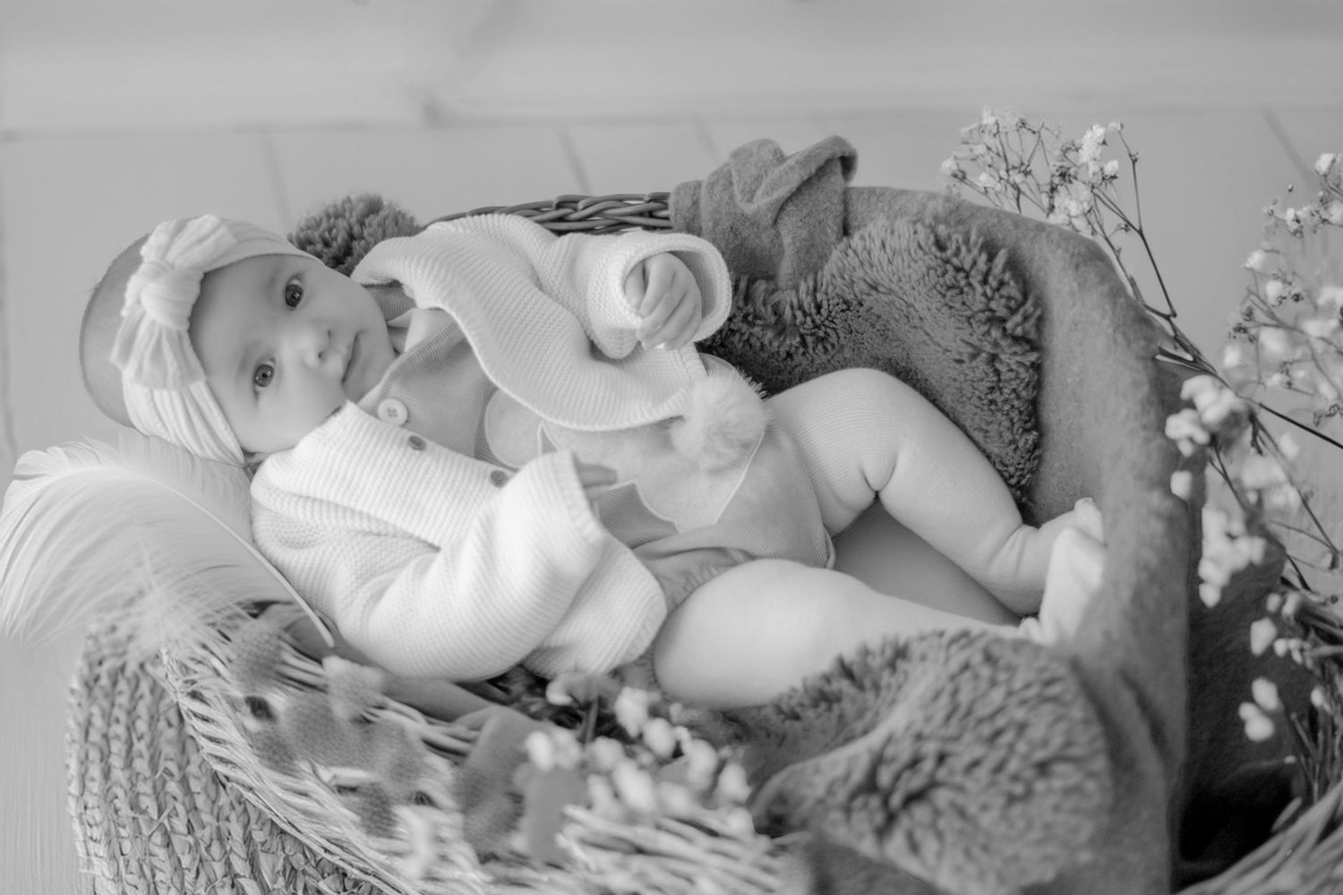 A baby lying in a woven basket wearing a white outfit next to delicate white flowers in black and white photography.
