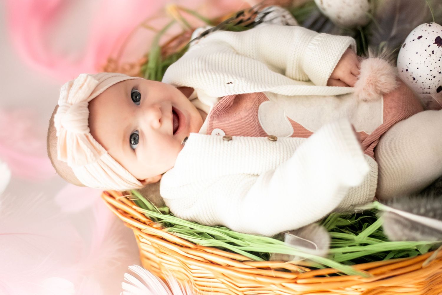 A small infant wearing a cream-colored outfit lies in a decorative wicker basket with green grass trim.