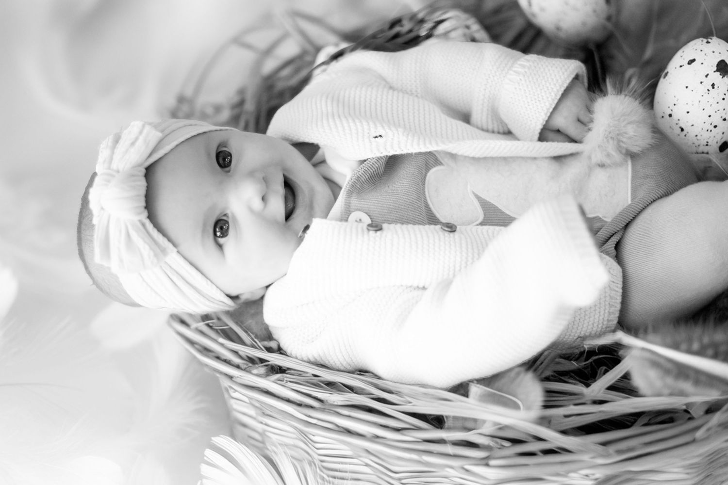 A black and white image of a baby laying in a wicker basket with soft blankets.
