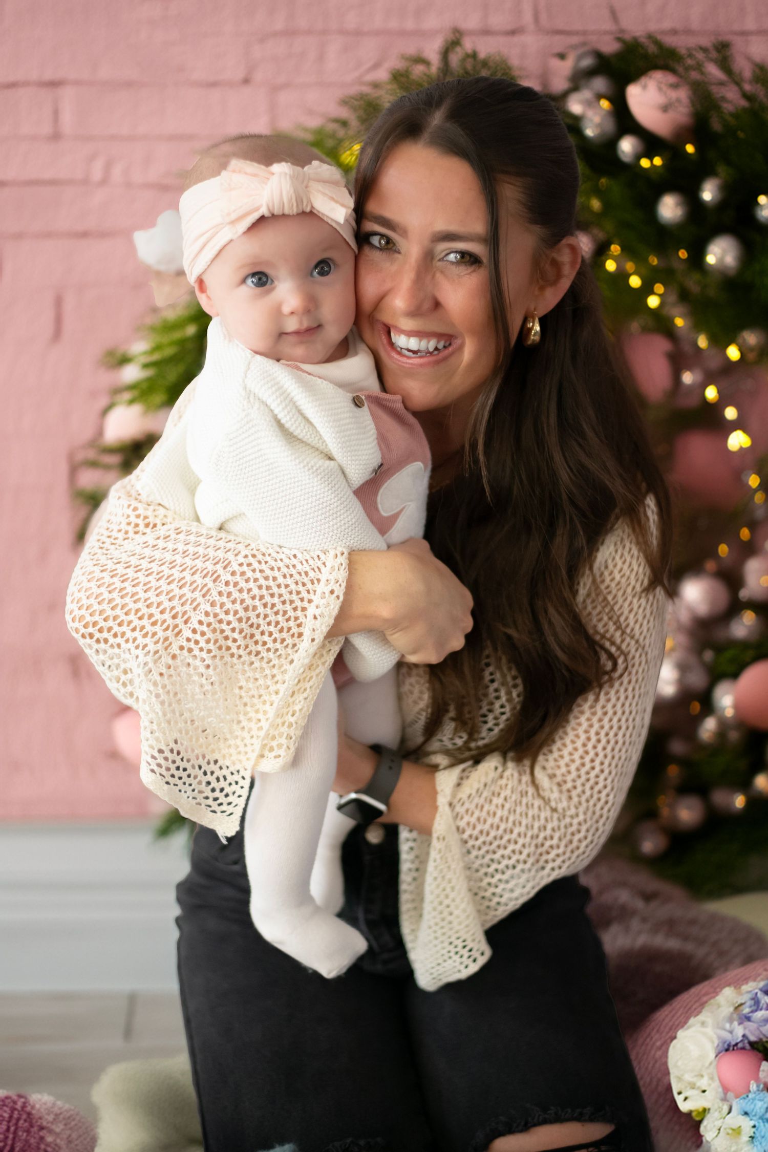 A festive holiday photo with a baby in white and a woman in a knit sweater in front of a Christmas tree.