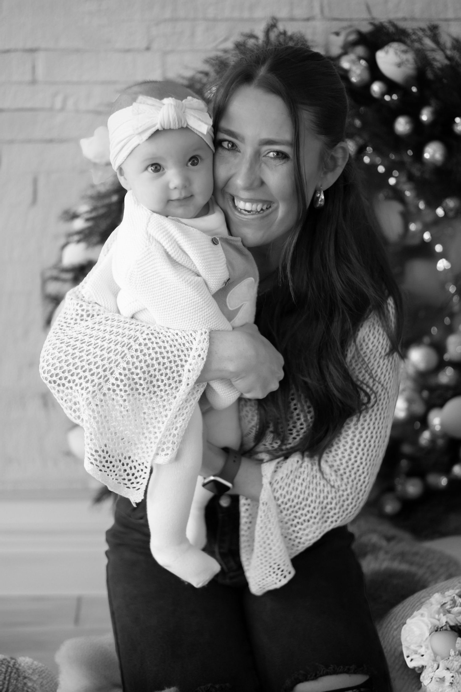 Black and white holiday portrait of adult wearing white sweater holding baby in lace dress by Christmas tree.