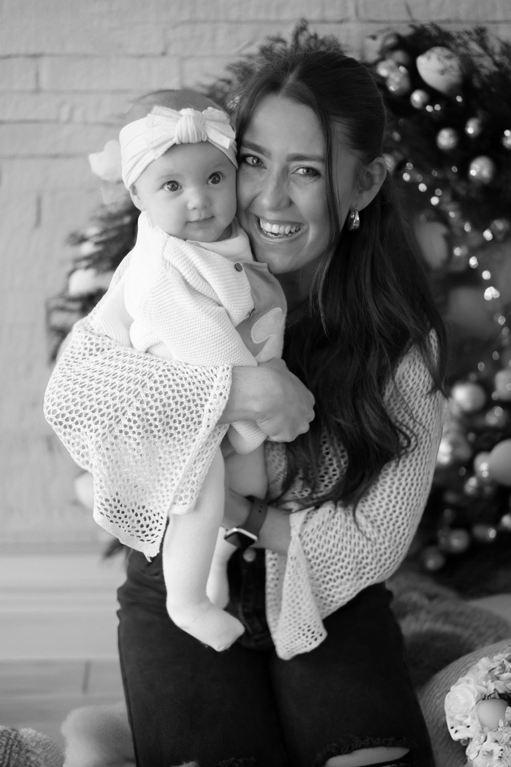 Black and white holiday portrait of adult wearing white sweater holding baby in lace dress by Christmas tree.