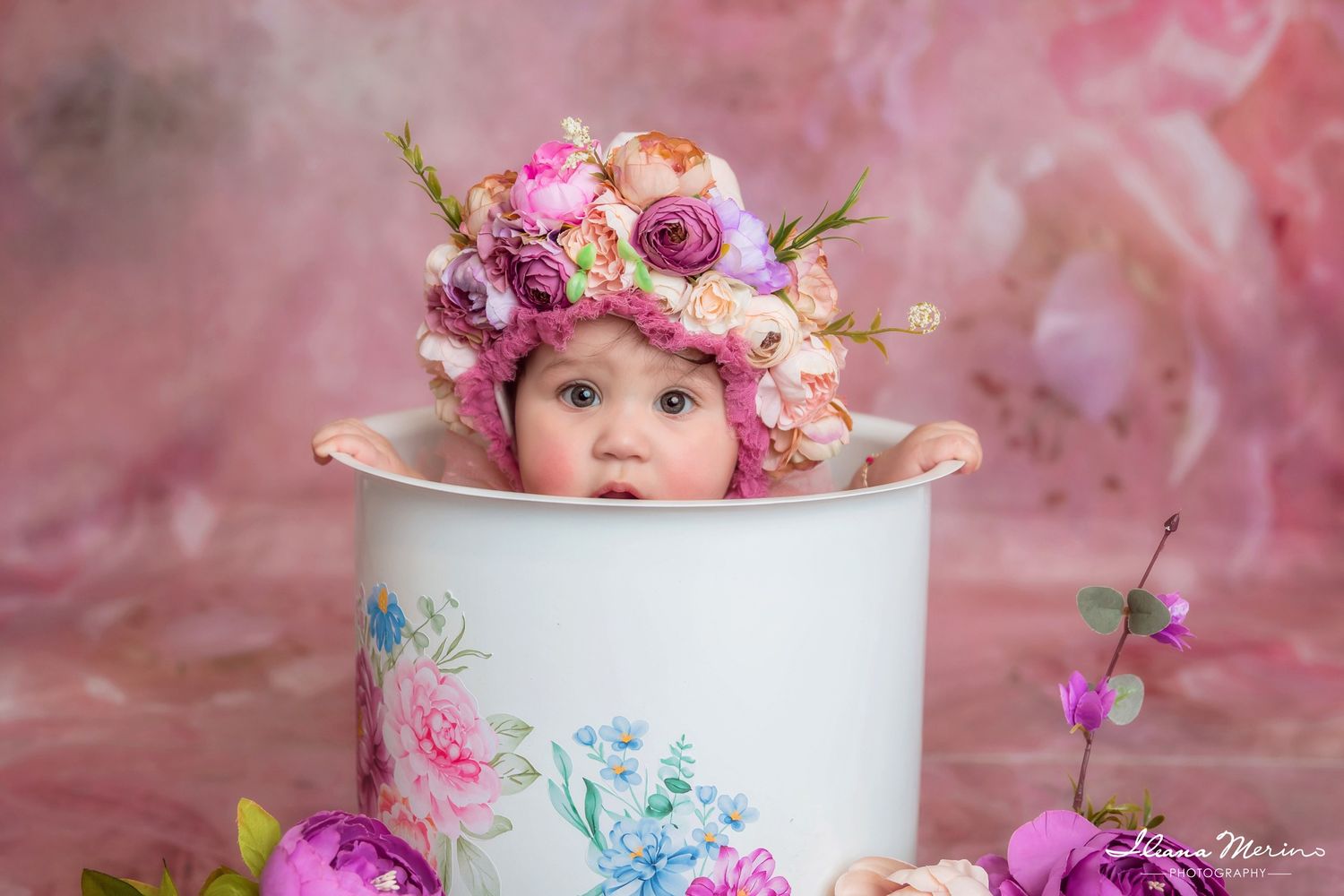 Adorable baby wearing a floral bonnet peeking out of a white floral bucket against a pink background.