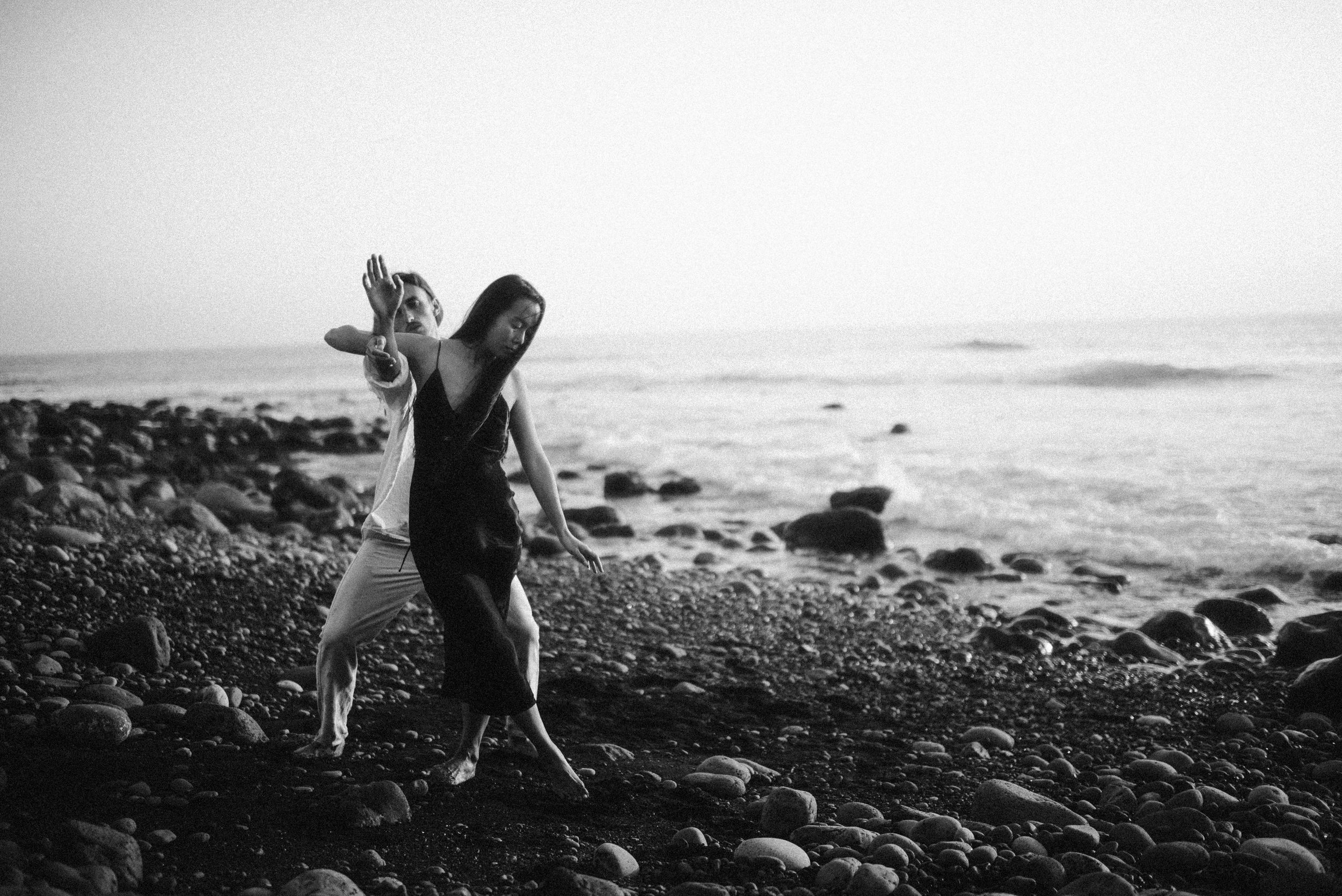 Black and white photo of someone walking on a rocky beach at the water's edge.