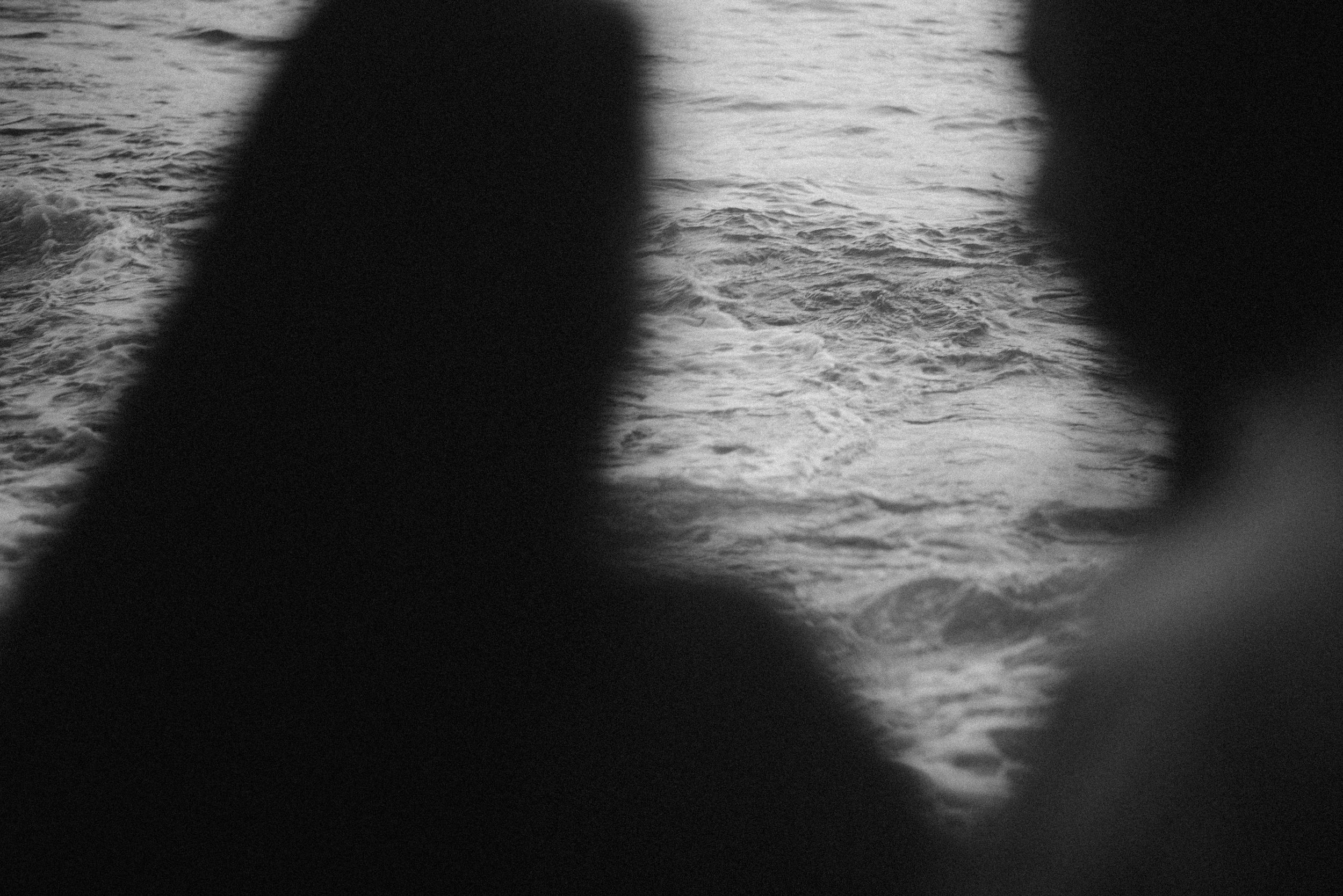 Two dark silhouettes are reflected in rippling water against a monochromatic background.