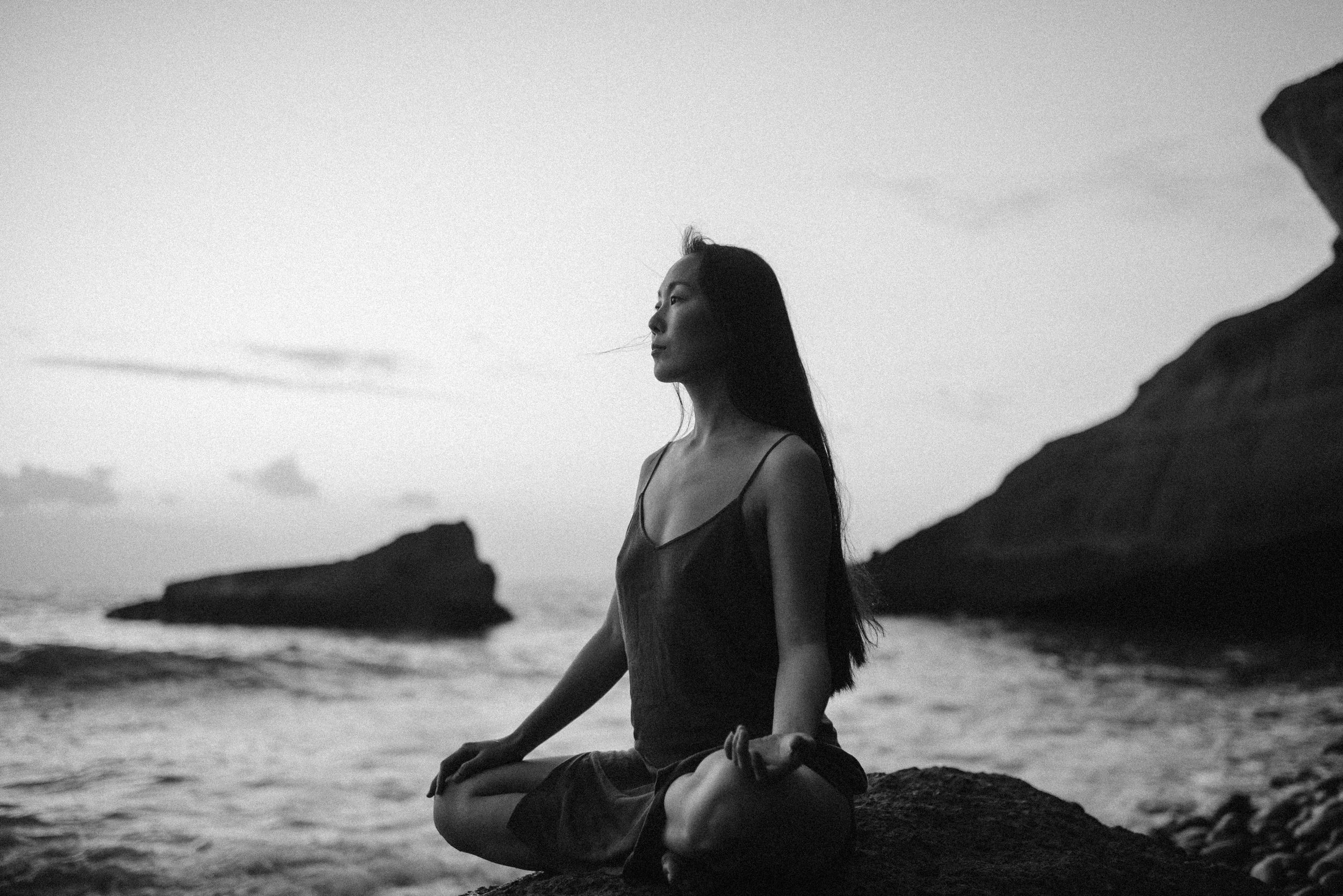Person meditating in lotus pose on rocky coastline at sunset in dramatic black and white composition.