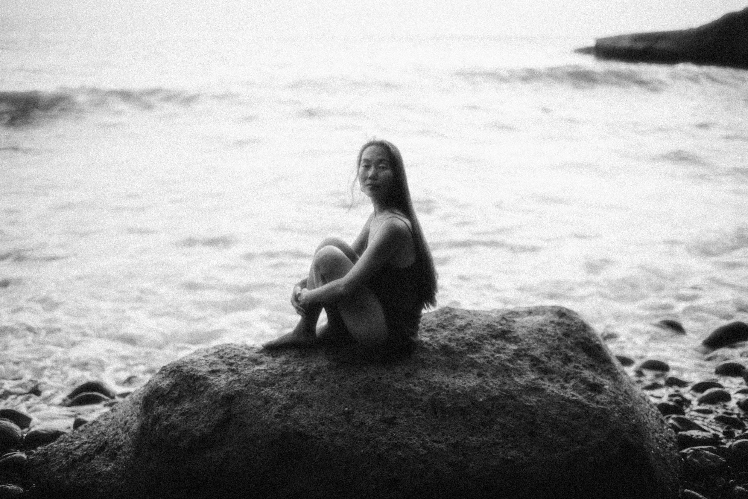 A silhouetted figure sitting alone on a rocky beach overlooking the ocean waves in black and white photography.