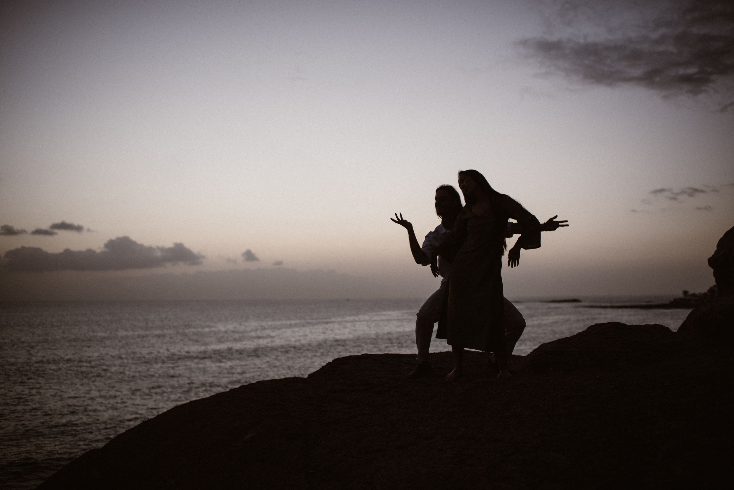 Silhouetted figures dance on a rocky cliff overlooking the ocean at sunset, creating a romantic and playful scene.