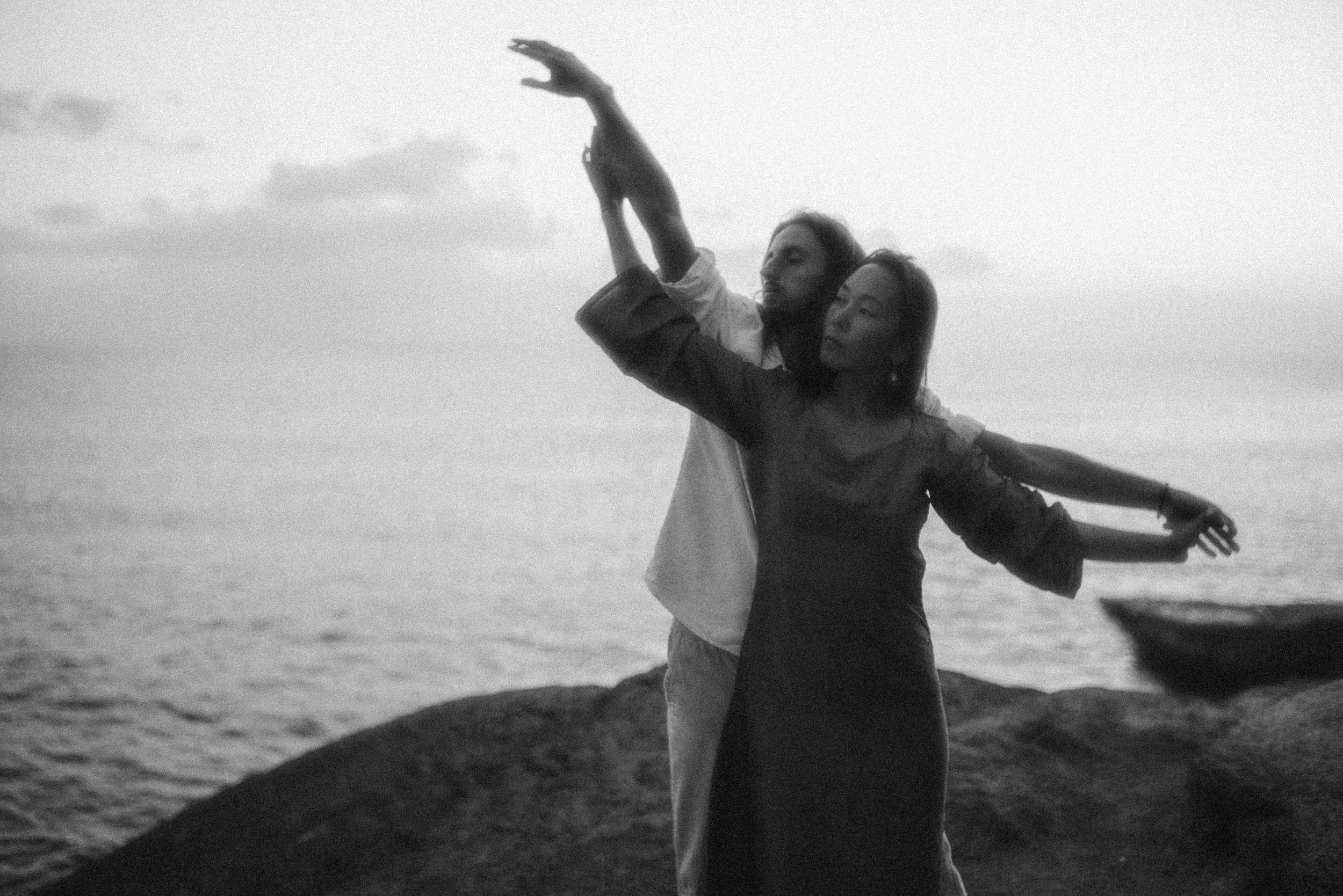 Two silhouettes dance together against an ocean backdrop on rocky shoreline.