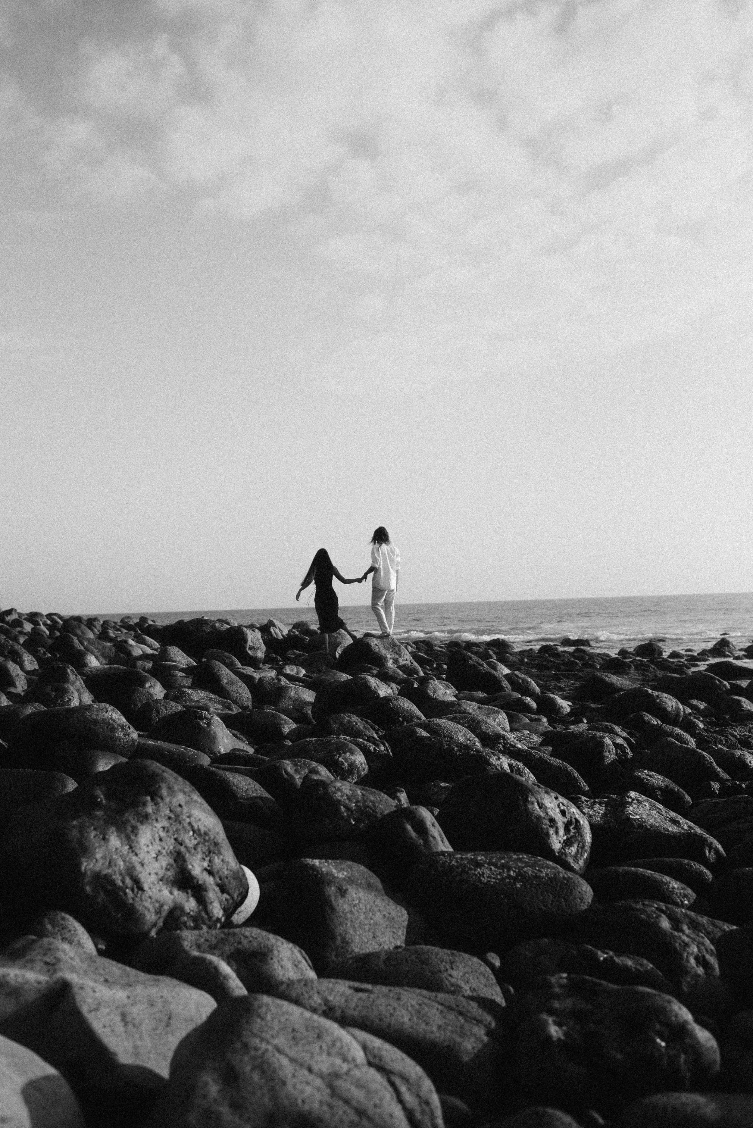 A black and white photograph of two silhouettes walking hand in hand across rocky coastal terrain.