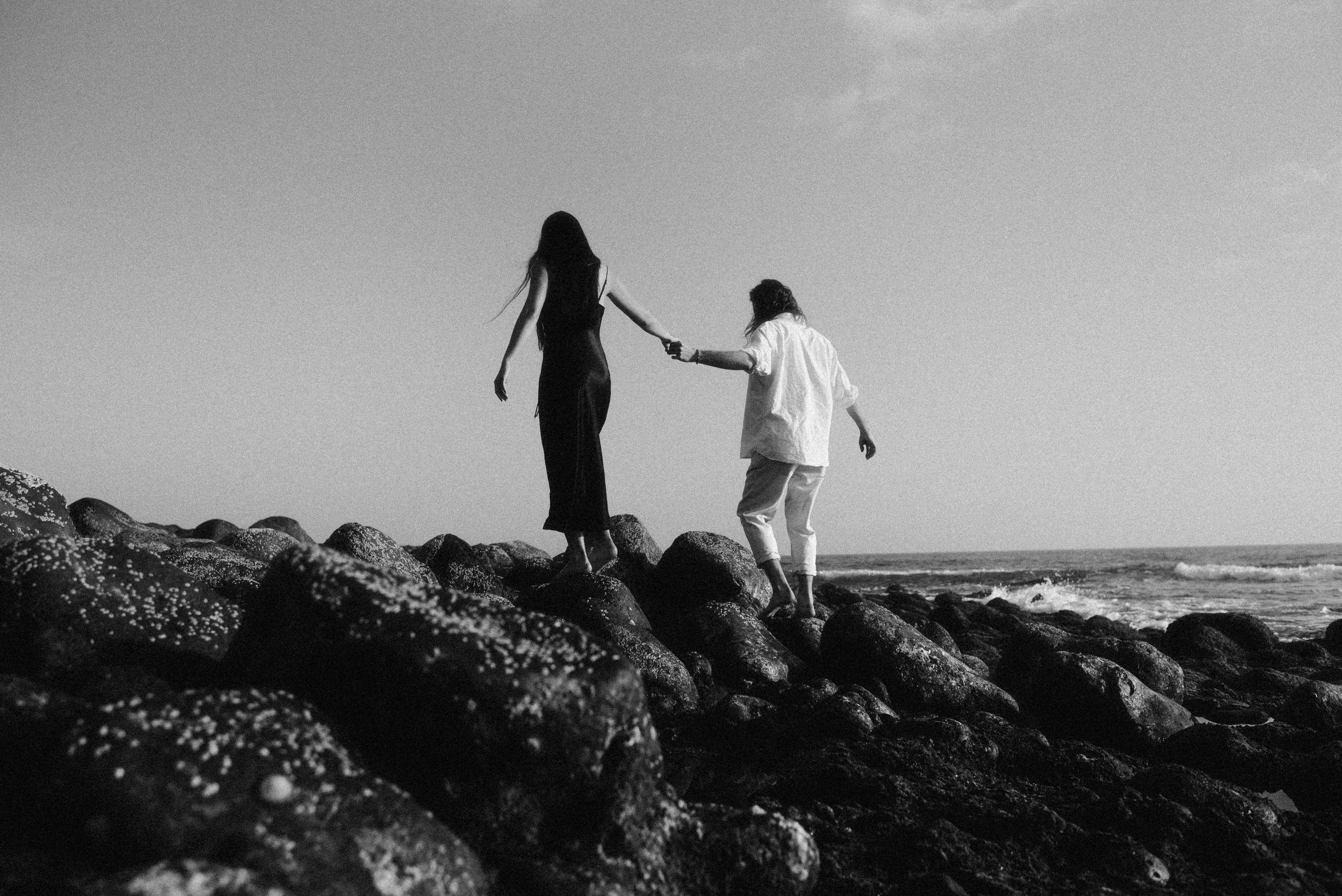 Two figures hold hands while walking along rocky coastal cliffs with ocean waves in the background.