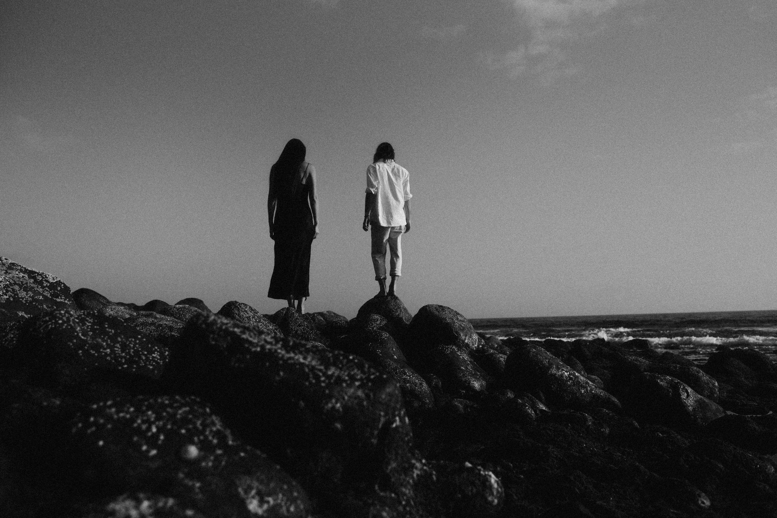Two silhouetted figures stand on rocky coastal outcrop against cloudy sky and ocean in black and white photography.
