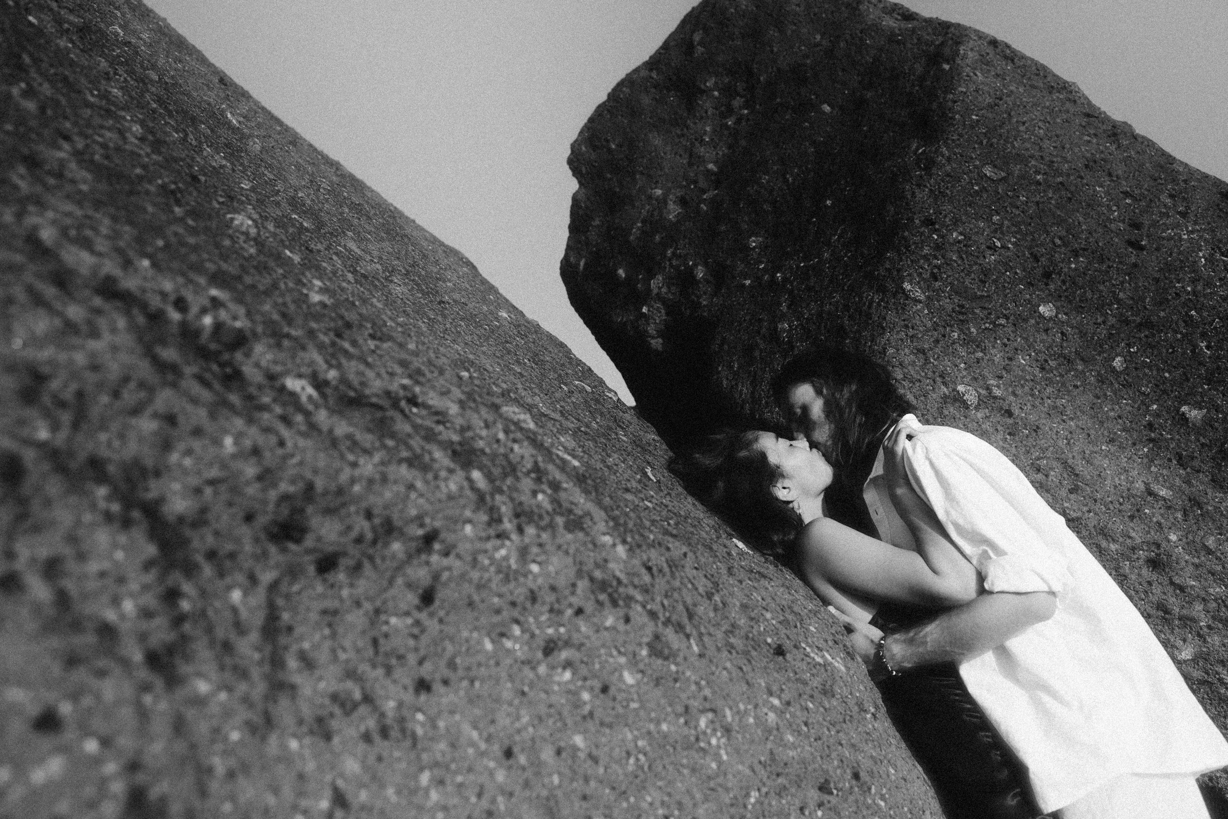 A person in white clothing peers curiously into a dark crevice between large boulders in a black and white photograph.