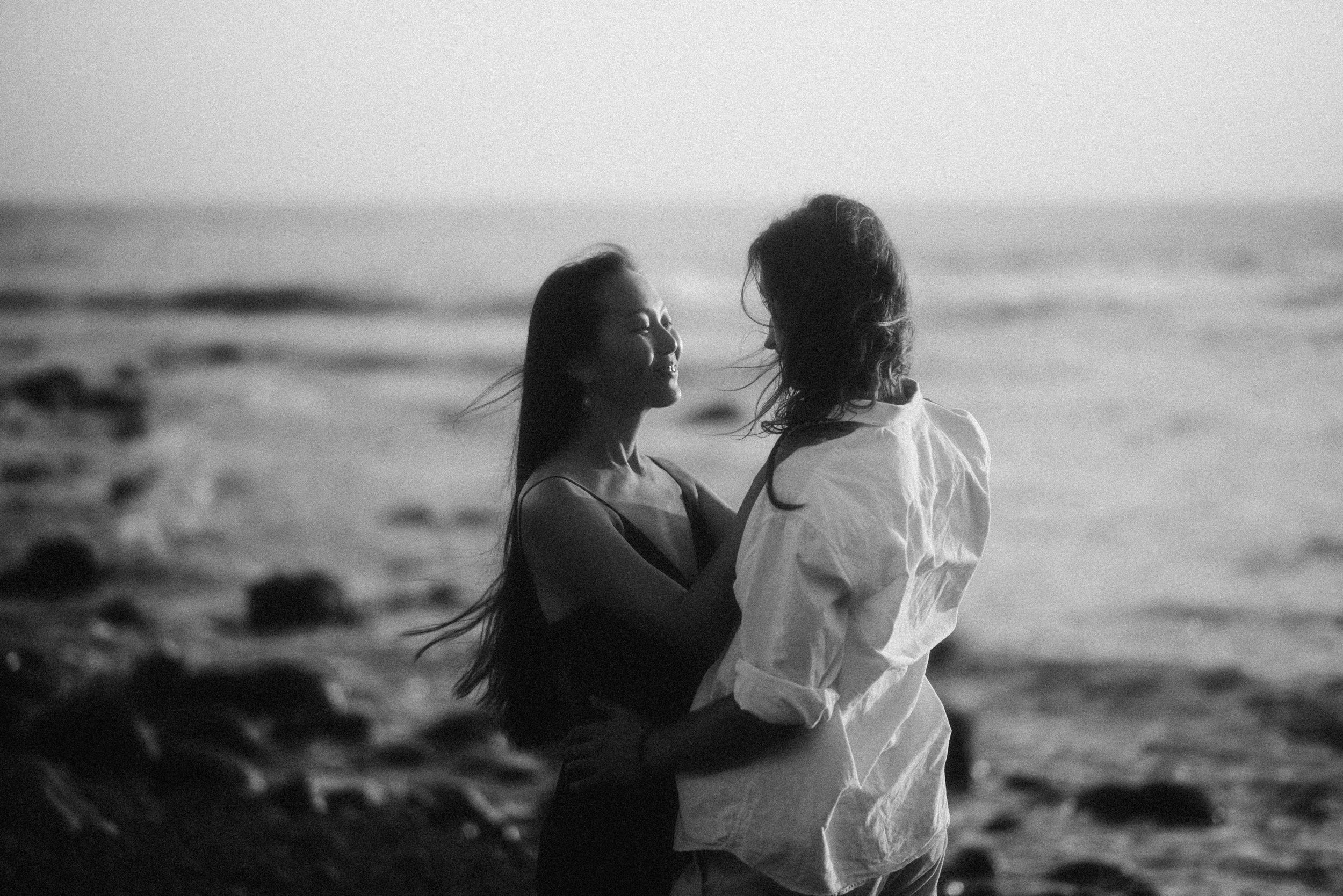 A romantic black and white silhouette of a couple embracing on a rocky beach at sunset with ocean waves in the background.