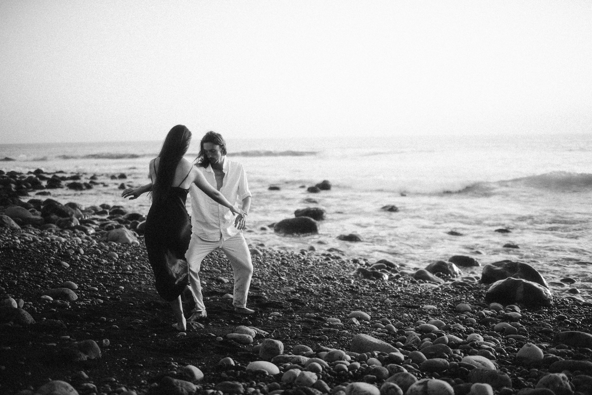 Silhouetted couple walking along a rocky beach at sunset with ocean waves in the background.