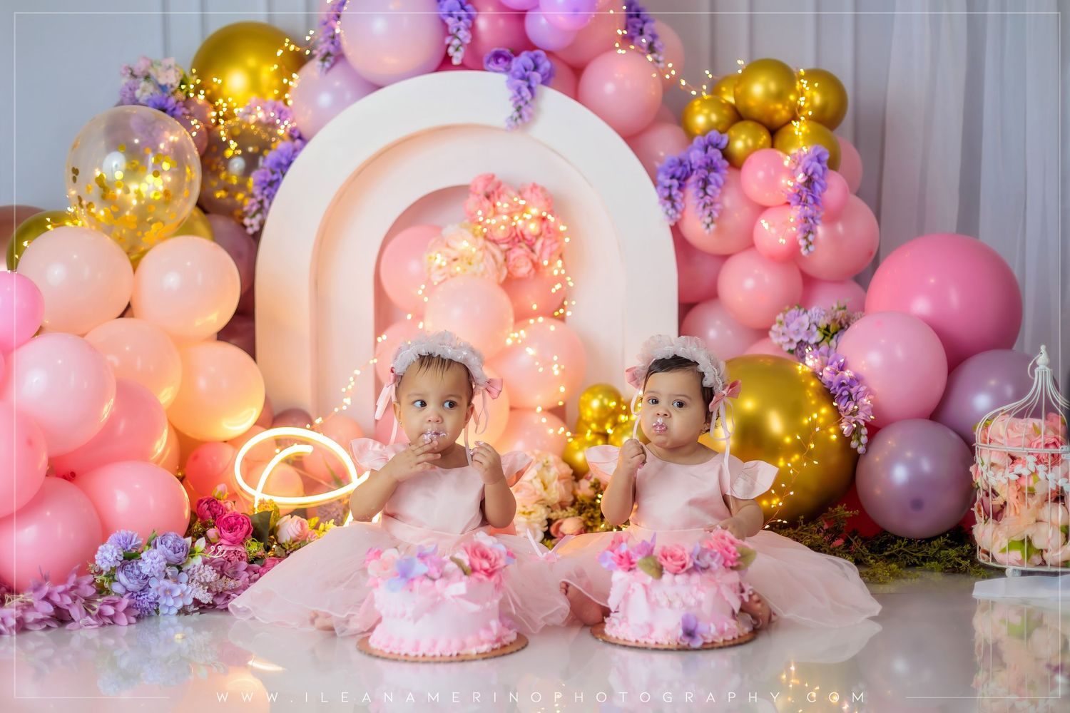 Twin baby girls in pink dresses celebrate their first birthday with cake smash amid colorful balloon decorations.