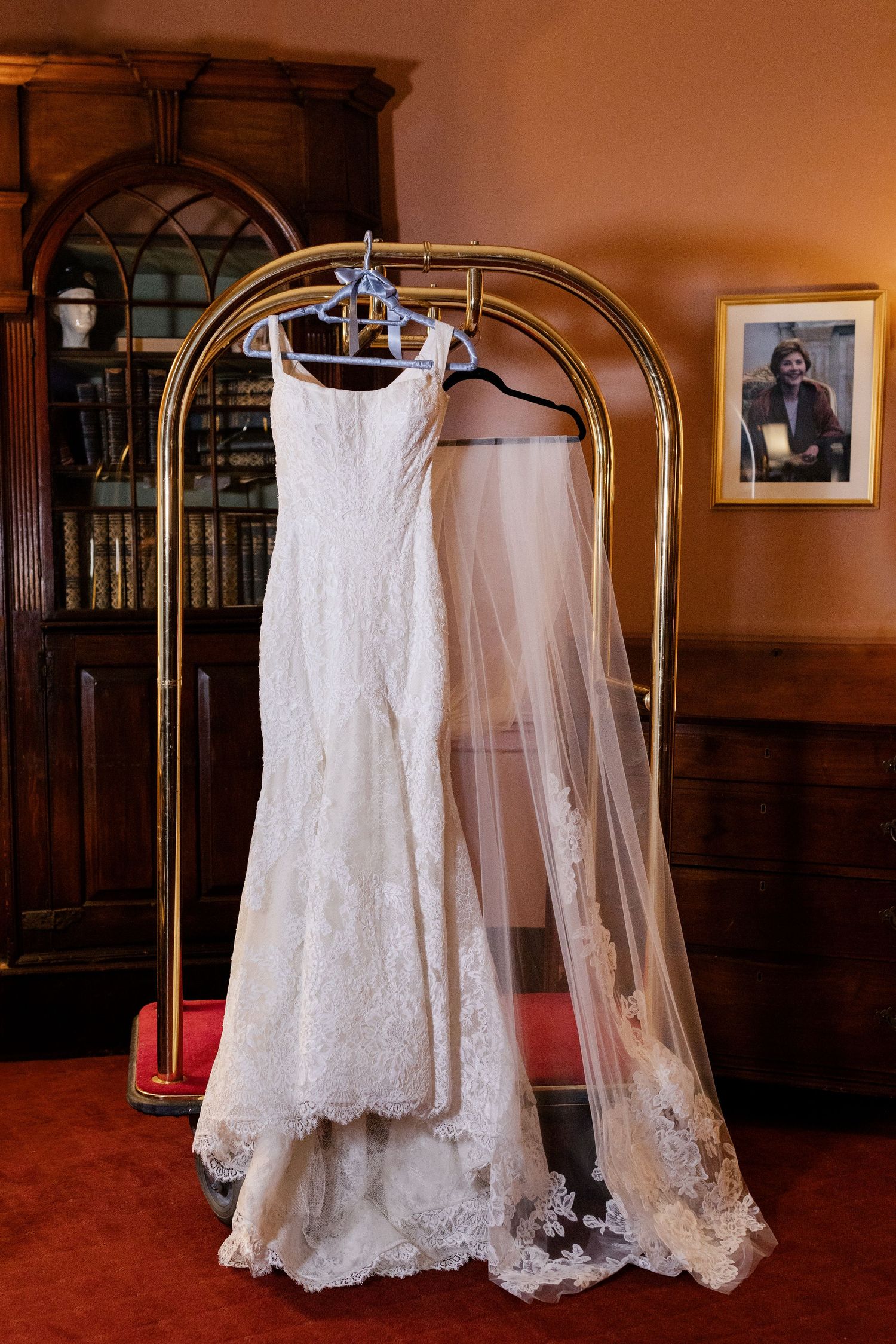 Elegant white lace wedding dress with train displayed on a brass garment rack against dark wood decor.