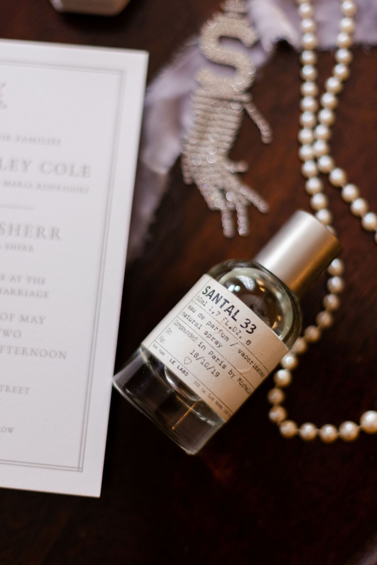 Pearl necklace and perfume bottle arranged with wedding stationery on dark wood surface.