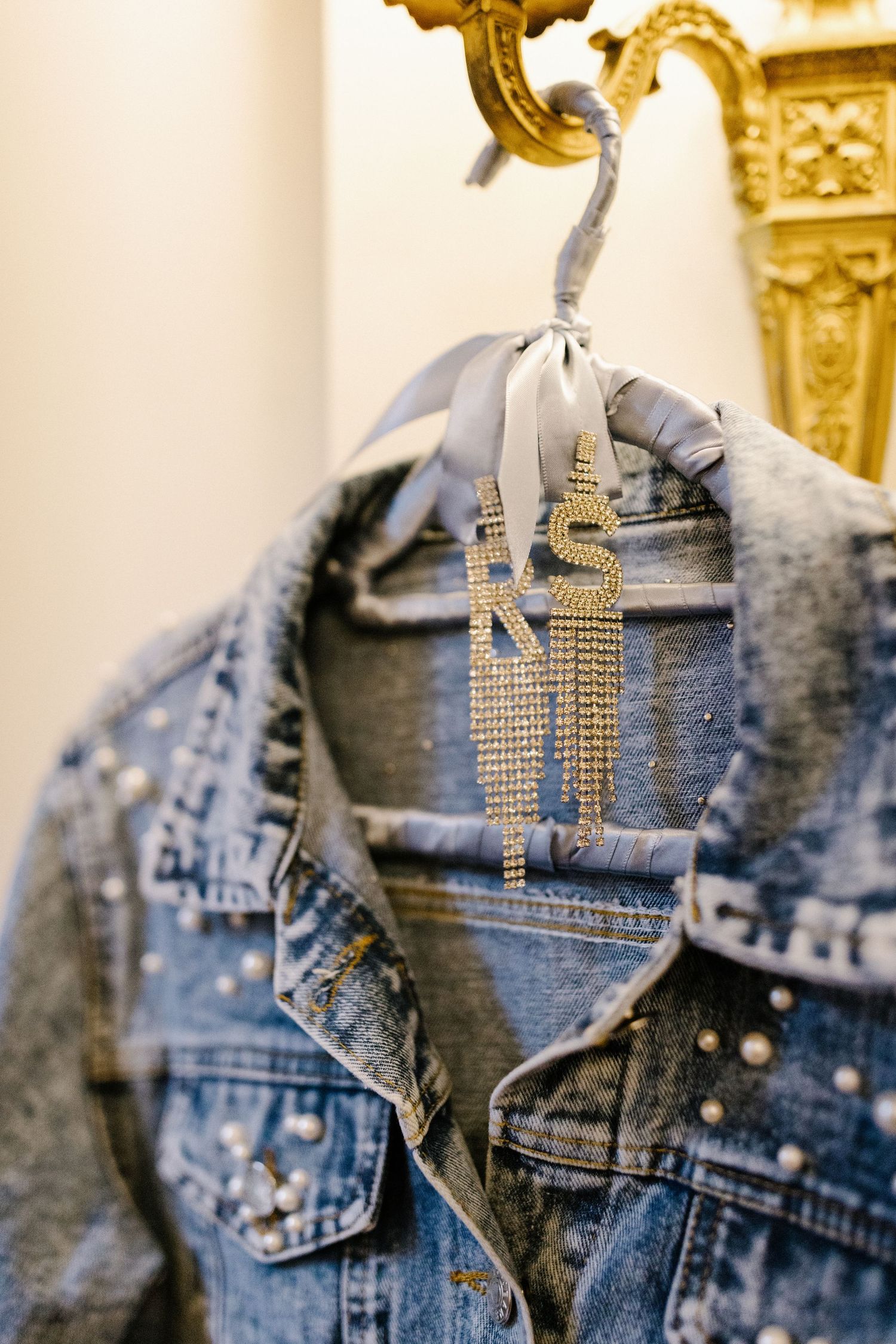 Elegant studded denim jacket hanging on decorative gold hanger against light background.