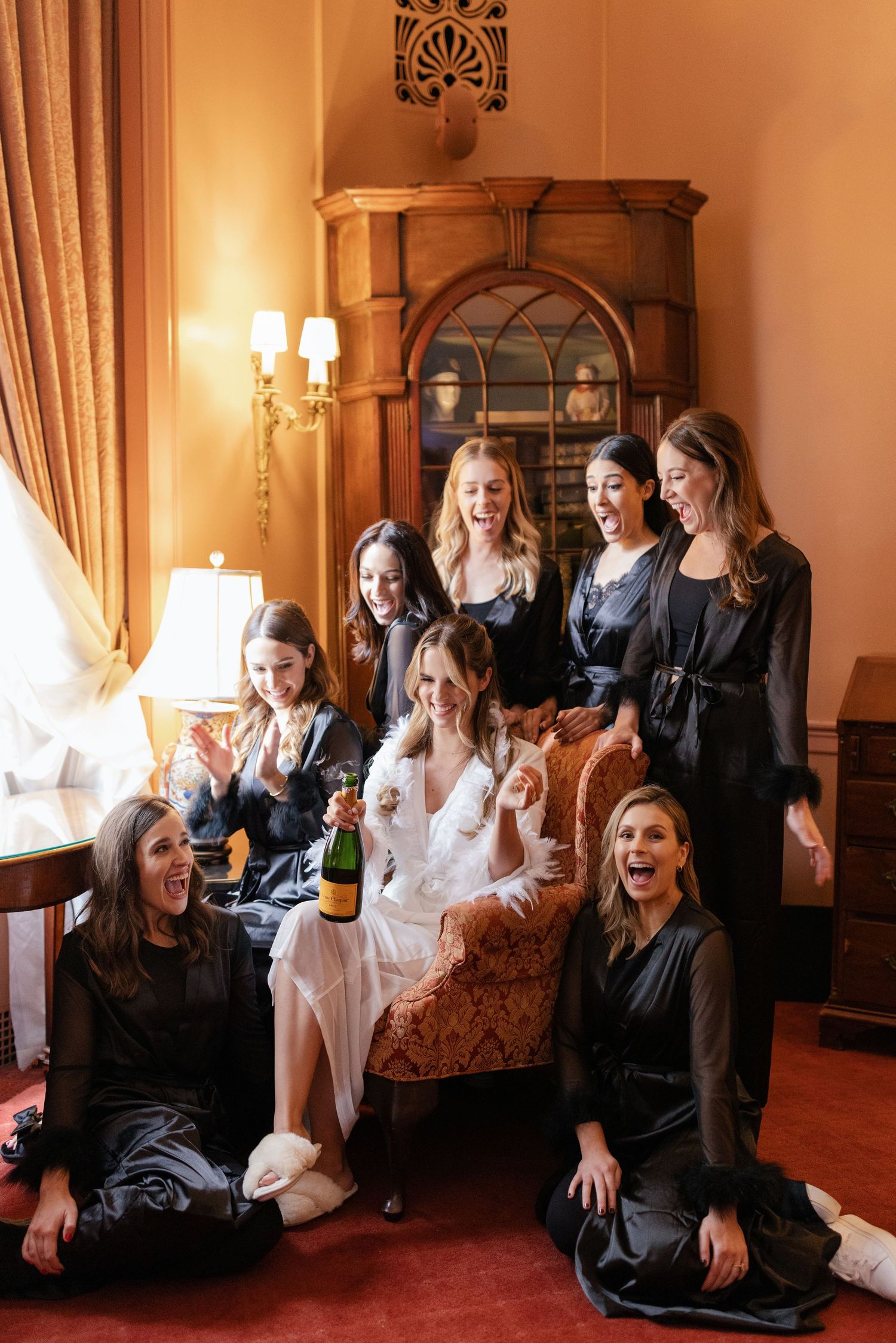 Bridal party in black dresses celebrates with champagne in an elegant room with warm lighting.