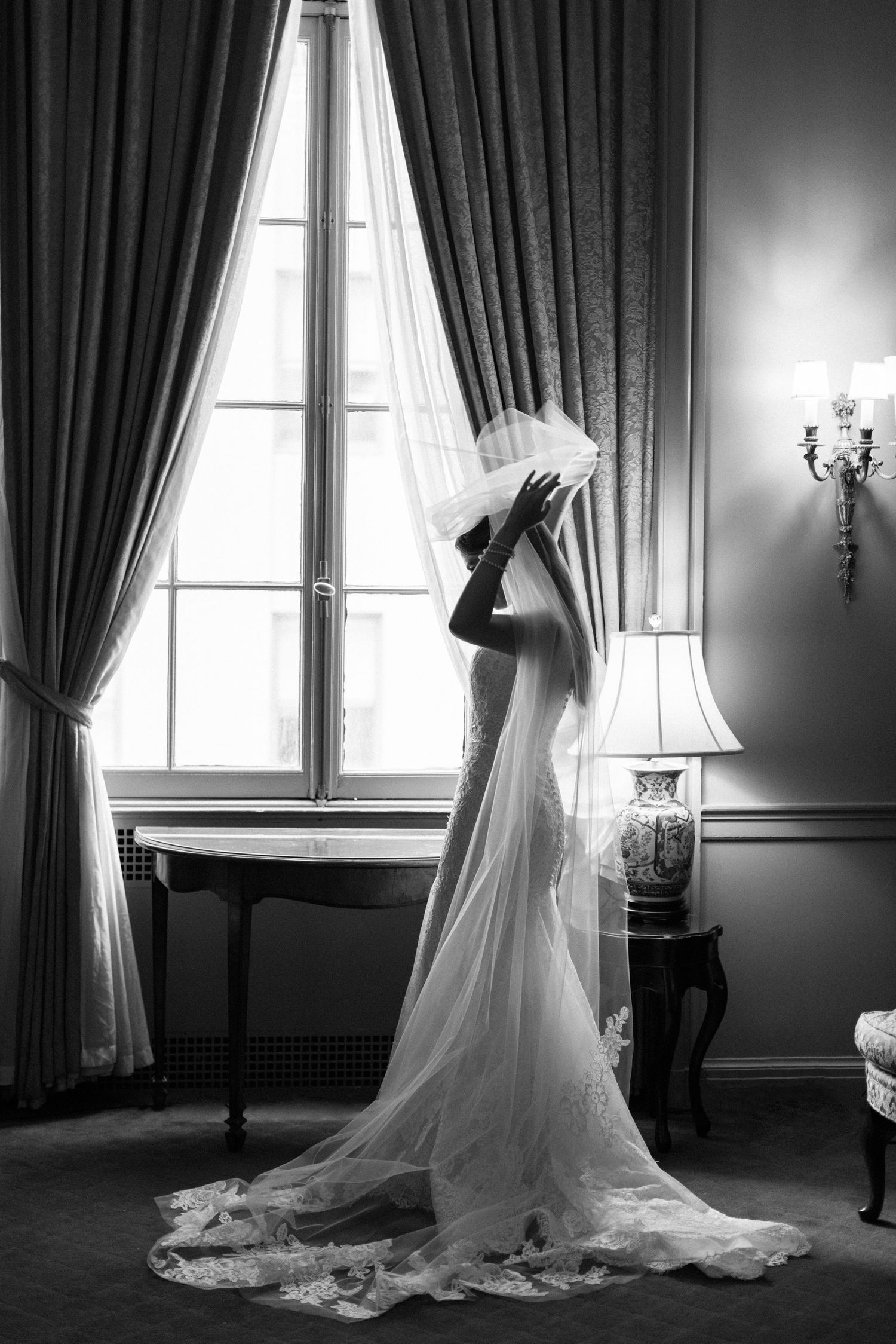 A bride in an elegant white wedding gown and veil poses dramatically by a tall window with floor-length curtains.