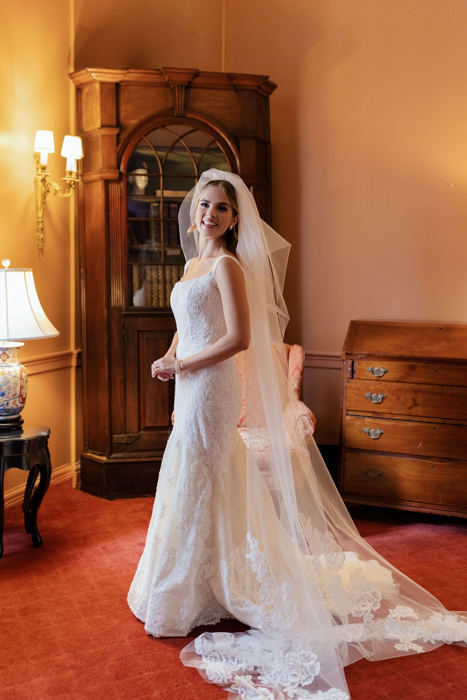 A bride in an elegant lace wedding dress and long veil poses in a warmly lit room with antique furniture.