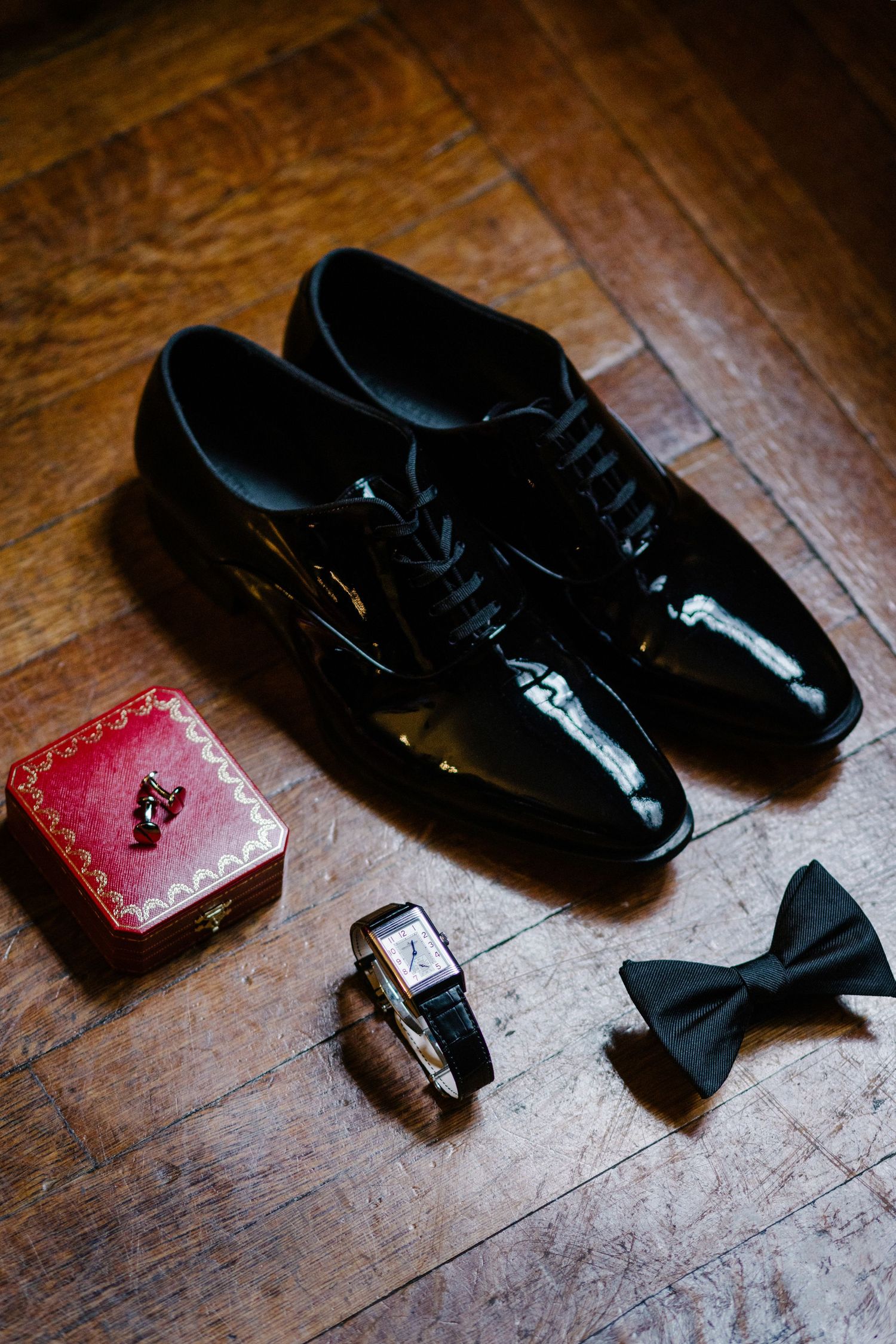 Wedding accessories including black patent leather dress shoes, bow tie, watch and red ring box on wooden floor.