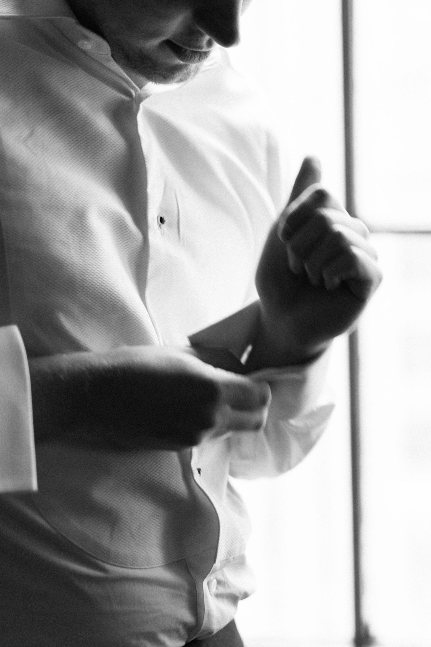 A groom adjusts his cufflinks in a black and white photo near a window.