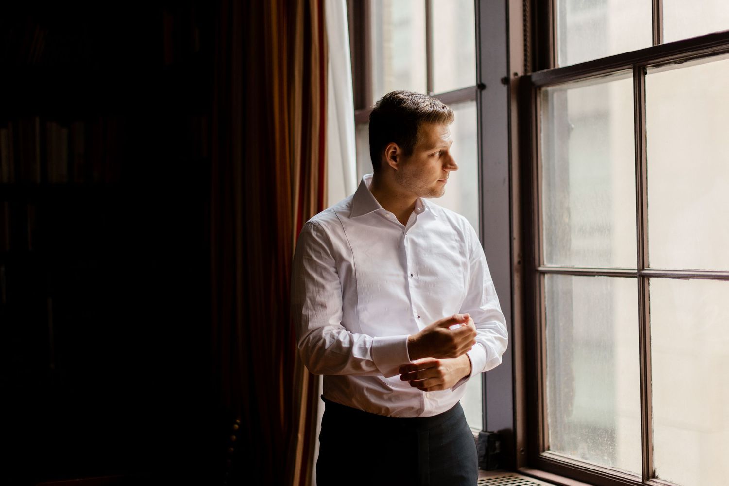 Professional in white button-down shirt stands contemplatively by window in natural lighting.