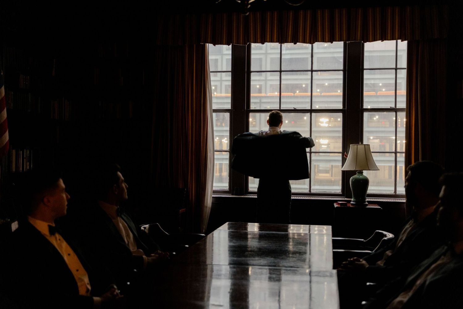 Silhouettes of people gathered in a dimly lit room with large windows and curtains, creating a moody atmosphere.