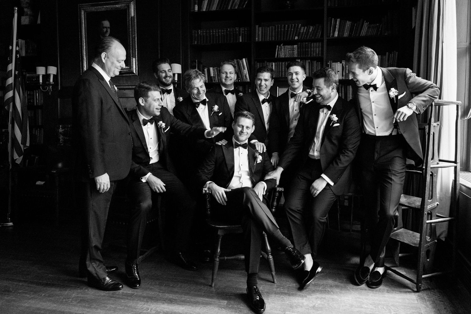 Groomsmen in tuxedos pose for a fun black and white portrait in a formal interior setting.